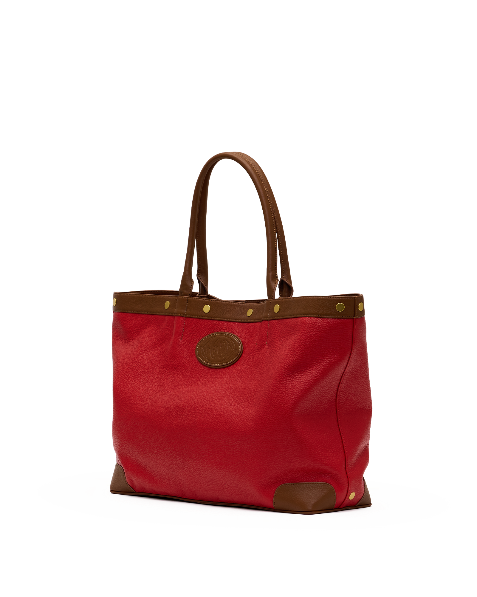 A red tote bag with brown leather handles, trim, and corner protectors, featuring brass-tone hardware and a circular brown logo patch on the front, set against a plain white background.