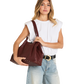 A woman modeling a large burgundy leather shoulder bag with studded details, styled with a white sleeveless top, light blue jeans, and a black belt with a gold buckle, against a plain white background.