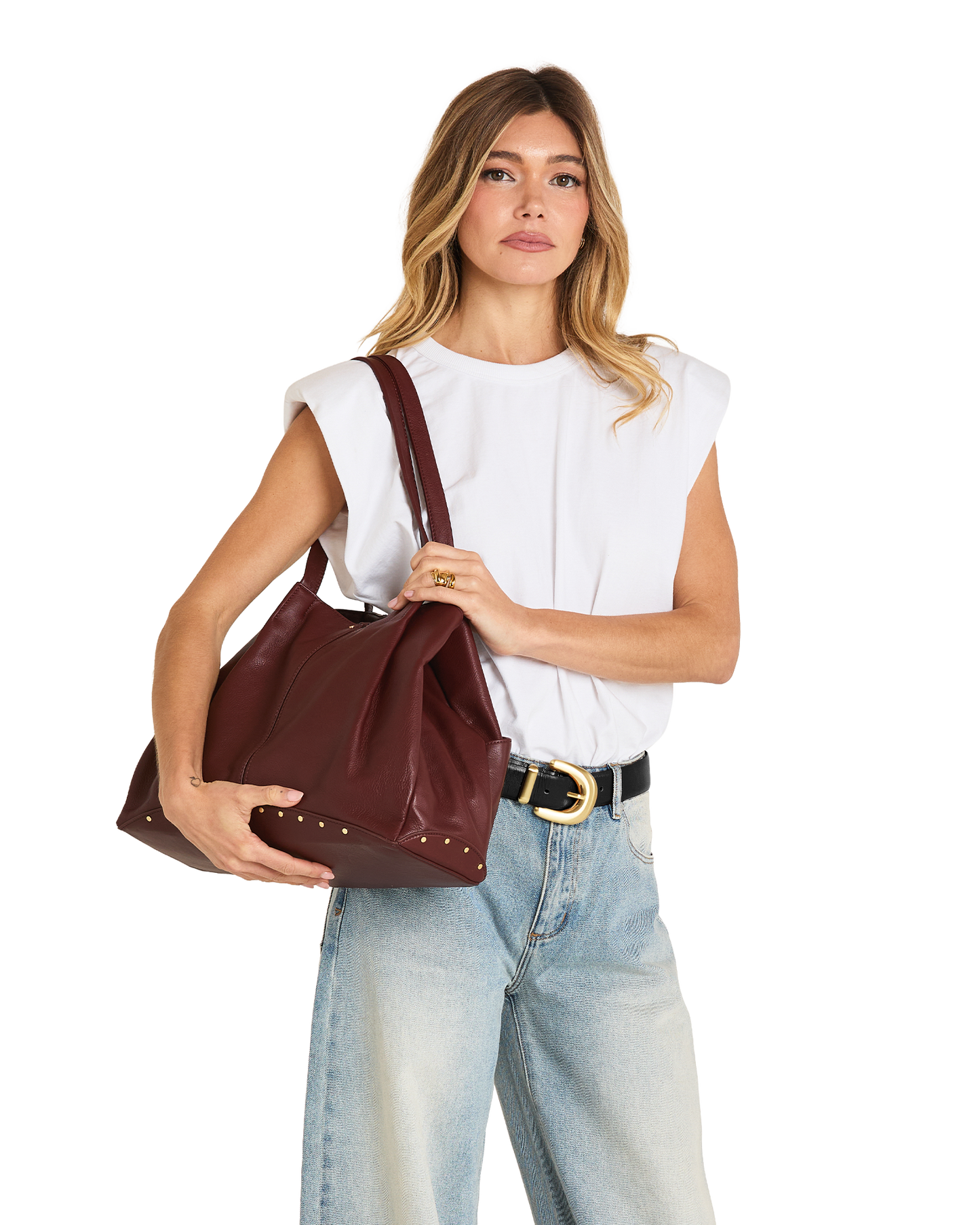 A woman modeling a large burgundy leather shoulder bag with studded details, styled with a white sleeveless top, light blue jeans, and a black belt with a gold buckle, against a plain white background.