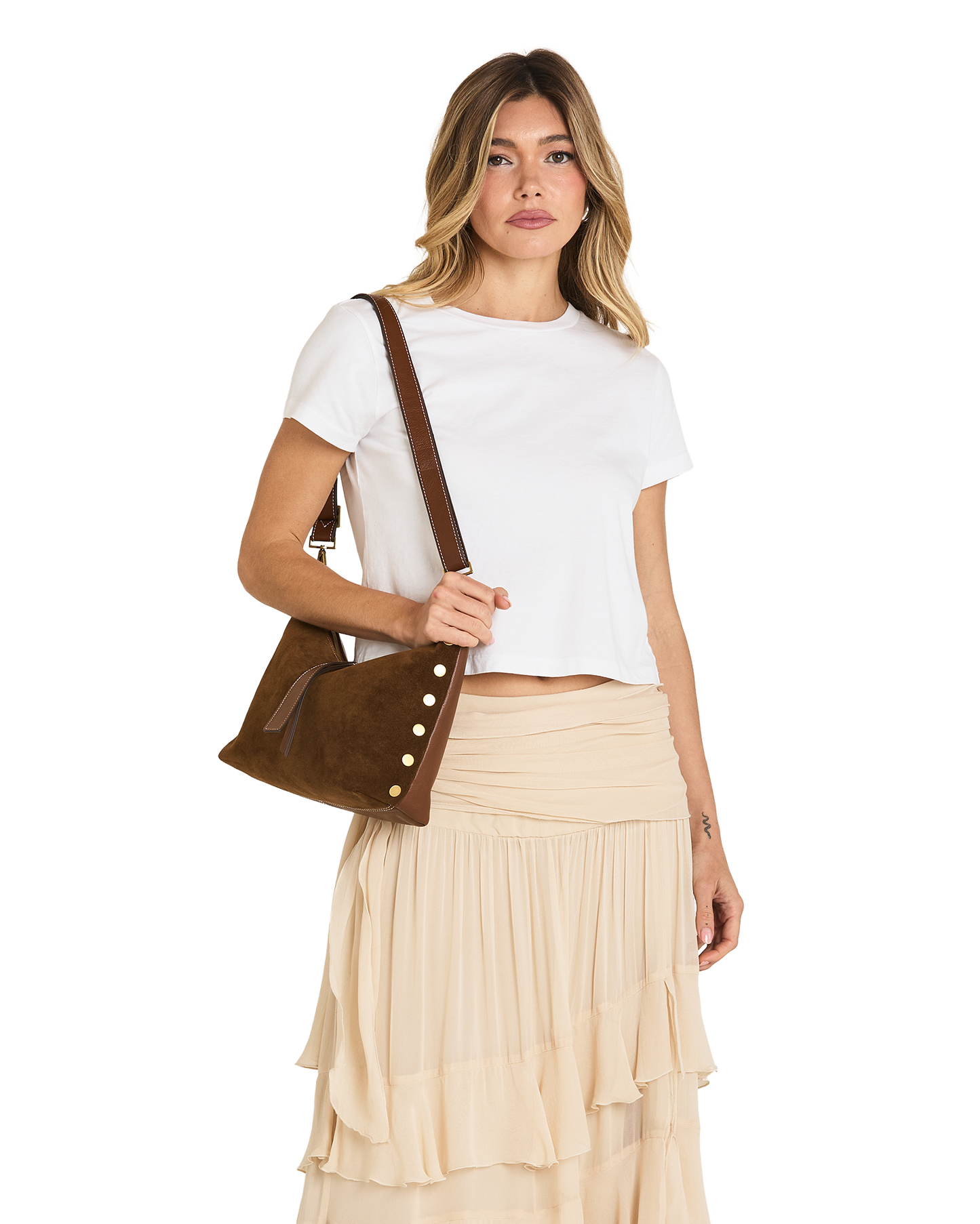 A woman modeling a brown suede crossbody bag with decorative gold-tone buttons, styled with a white crop top and a beige pleated skirt against a plain white background.