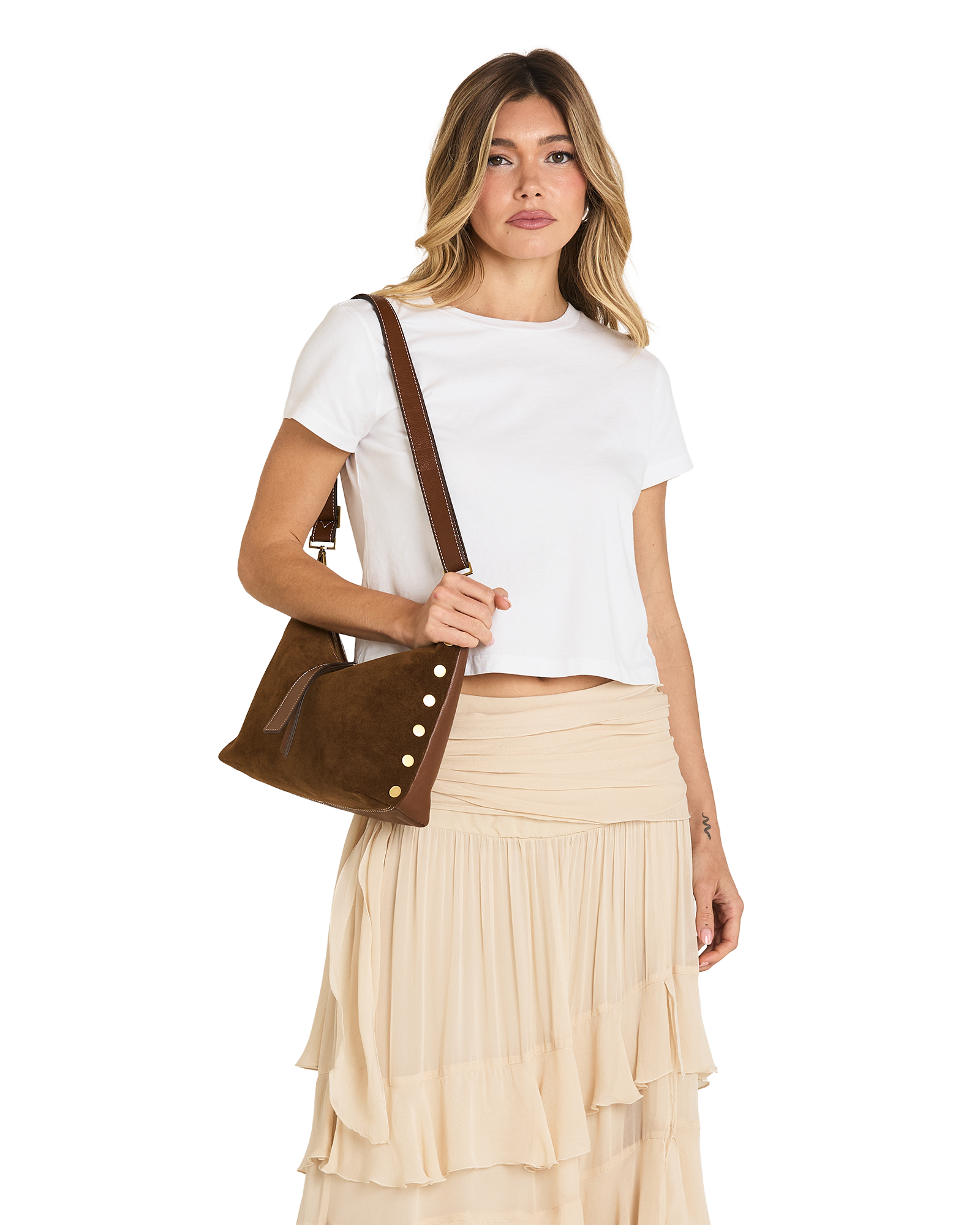 A woman modeling a brown suede crossbody bag with decorative gold-tone buttons, styled with a white crop top and a beige pleated skirt against a plain white background.