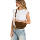 A woman wearing a white sleeveless top and light blue jeans models a brown suede crescent-shaped crossbody bag with gold-tone hardware against a plain white background.