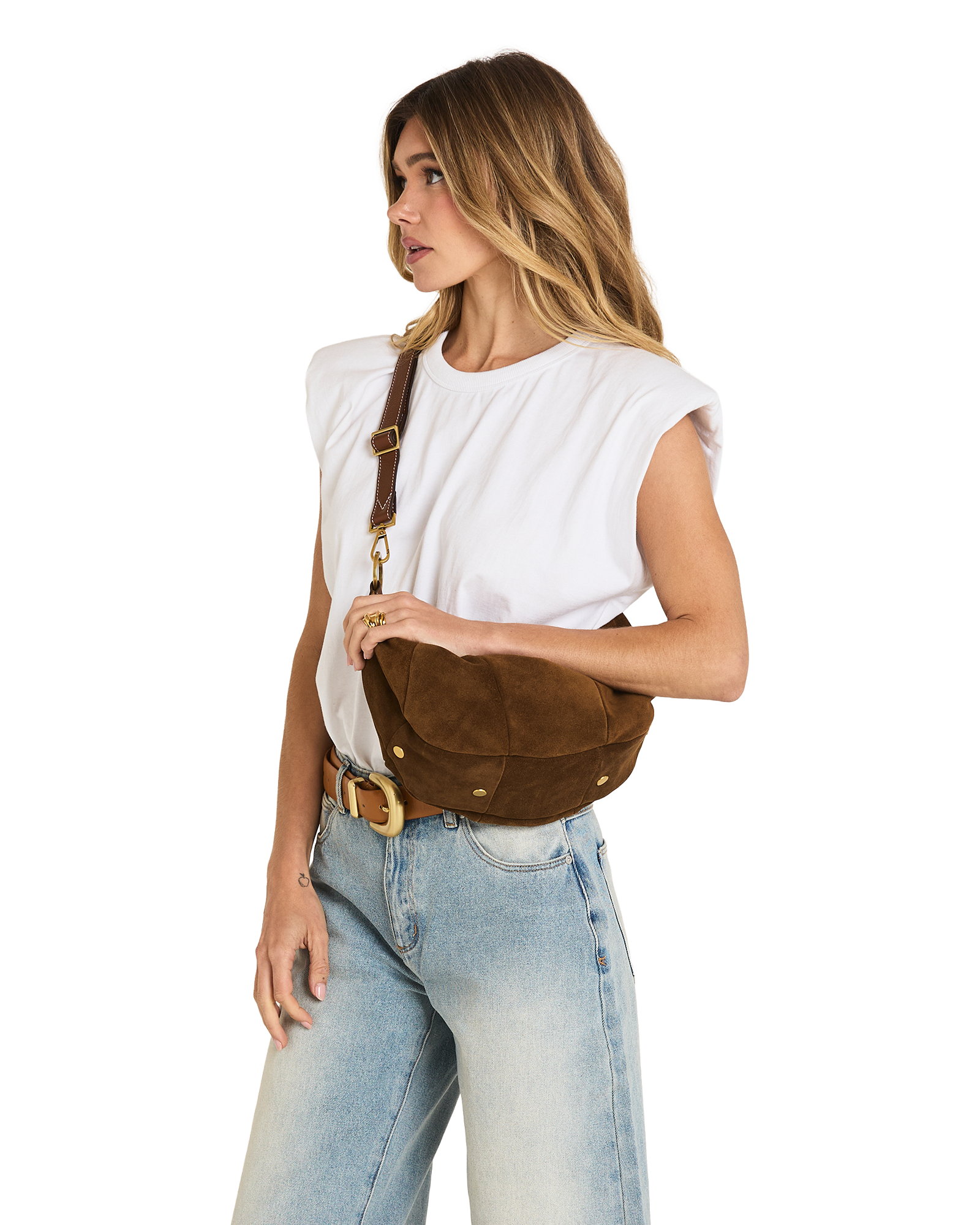 A woman wearing a white sleeveless top and light blue jeans models a brown suede crescent-shaped crossbody bag with gold-tone hardware against a plain white background.