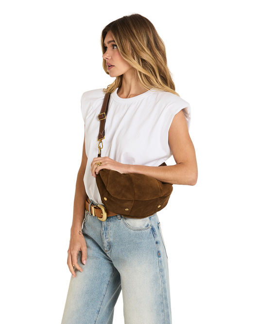 A woman wearing a white sleeveless top and light blue jeans models a brown suede crescent-shaped crossbody bag with gold-tone hardware against a plain white background.