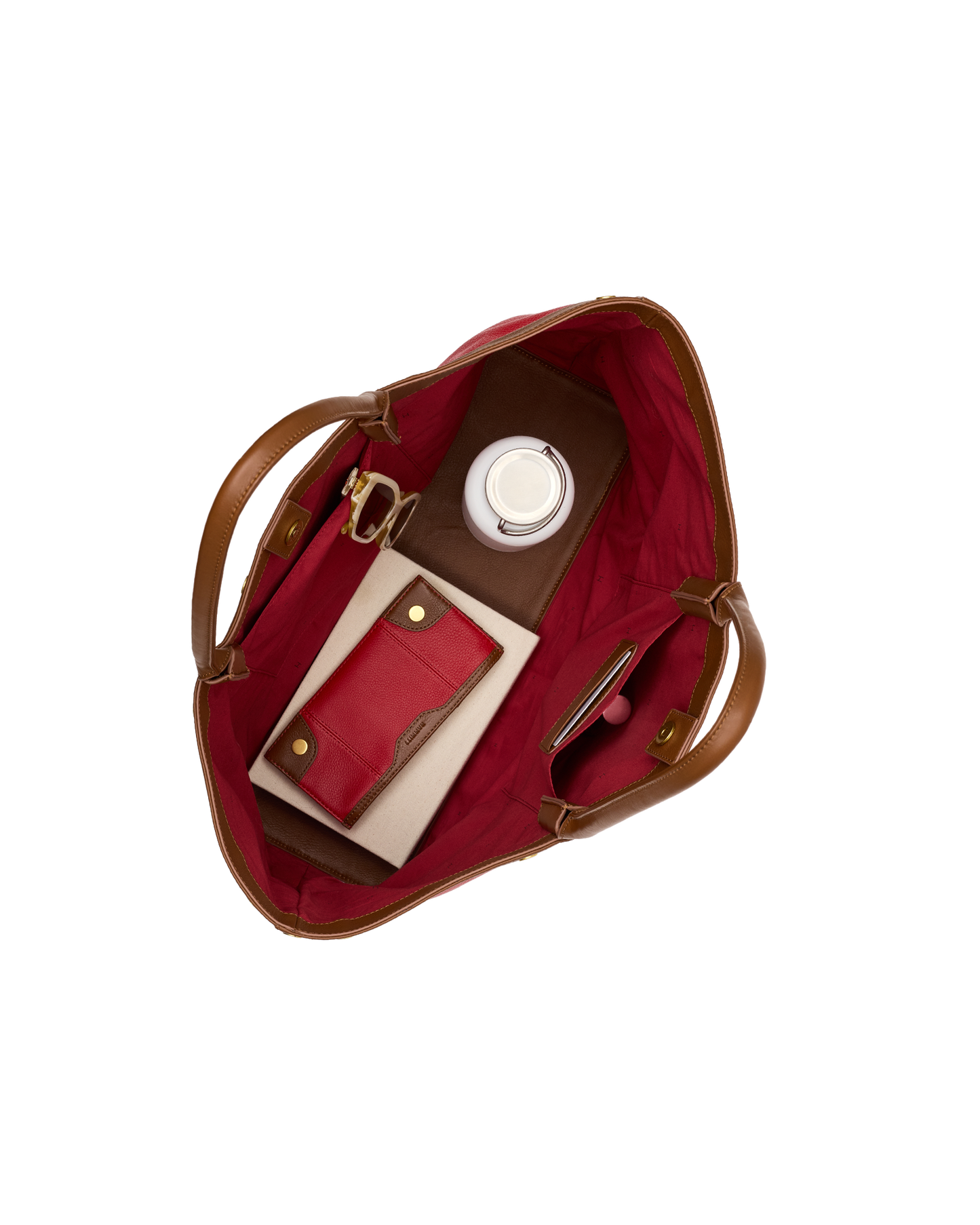 Top-down view of an open tote bag with a deep red interior and brown leather handles, shown filled with a matching red card holder, a brown leather wallet, a white circular item, and a slim pink accessory on a plain white background.