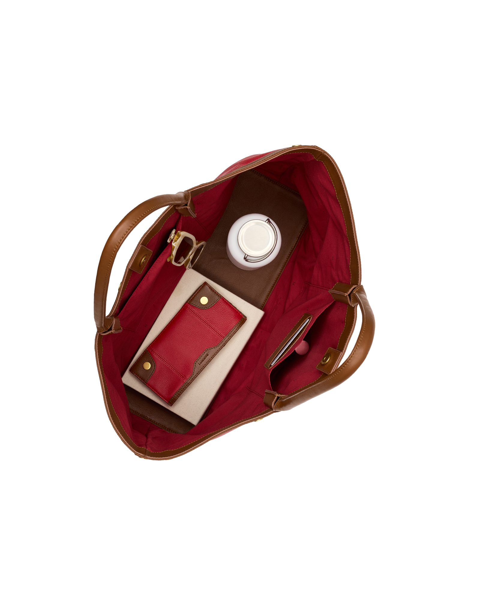 Top-down view of an open tote bag with a deep red interior and brown leather handles, shown filled with a matching red card holder, a brown leather wallet, a white circular item, and a slim pink accessory on a plain white background.