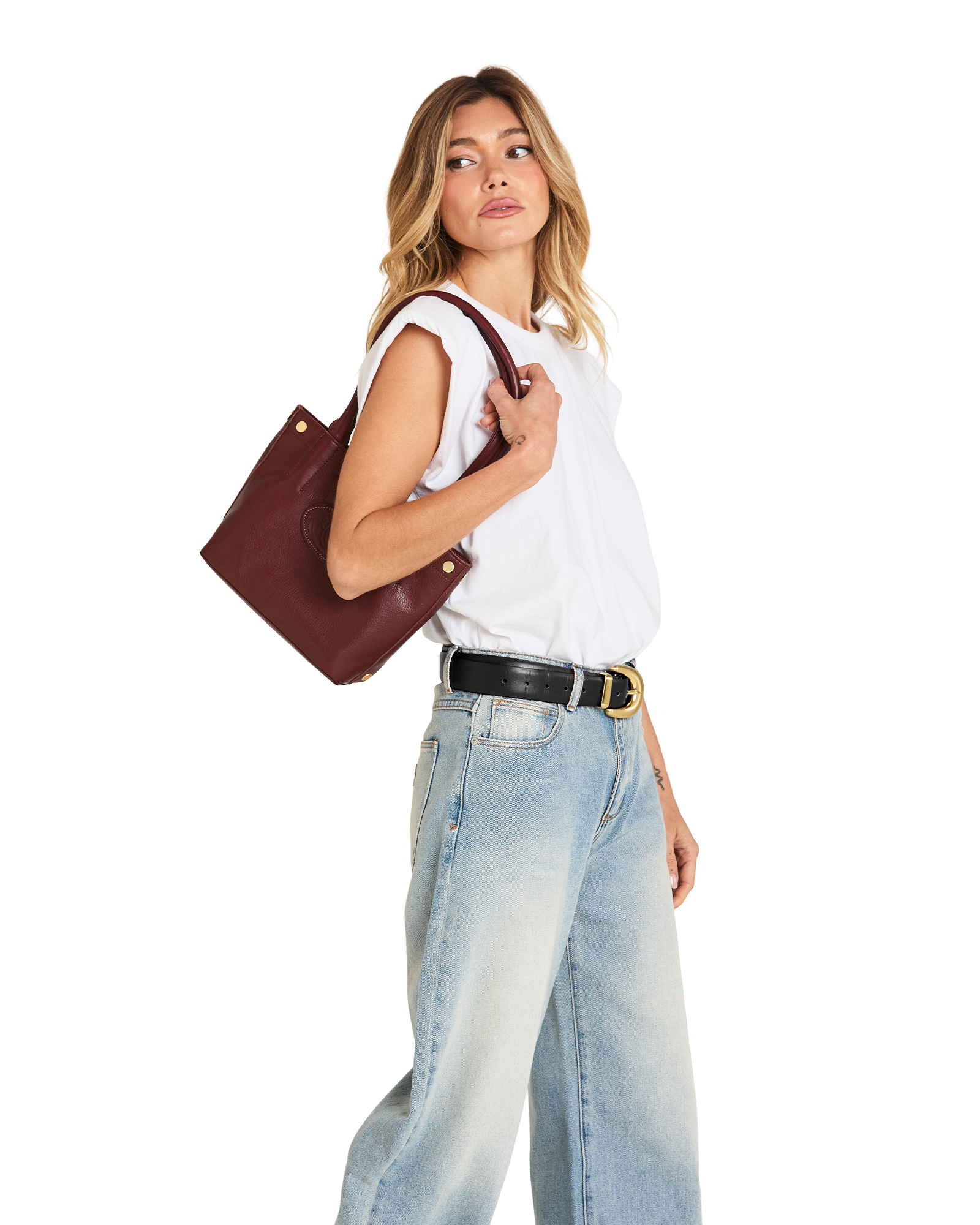 A woman wearing a white sleeveless top, light blue wide-leg jeans, and a black belt with a gold buckle, carrying a dark brown leather tote bag over her shoulder against a plain white background.