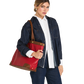 A woman modeling a large red leather tote bag with brown leather trim and gold-tone hardware, styled with a white button-down shirt and a dark denim jacket against a plain white background.
