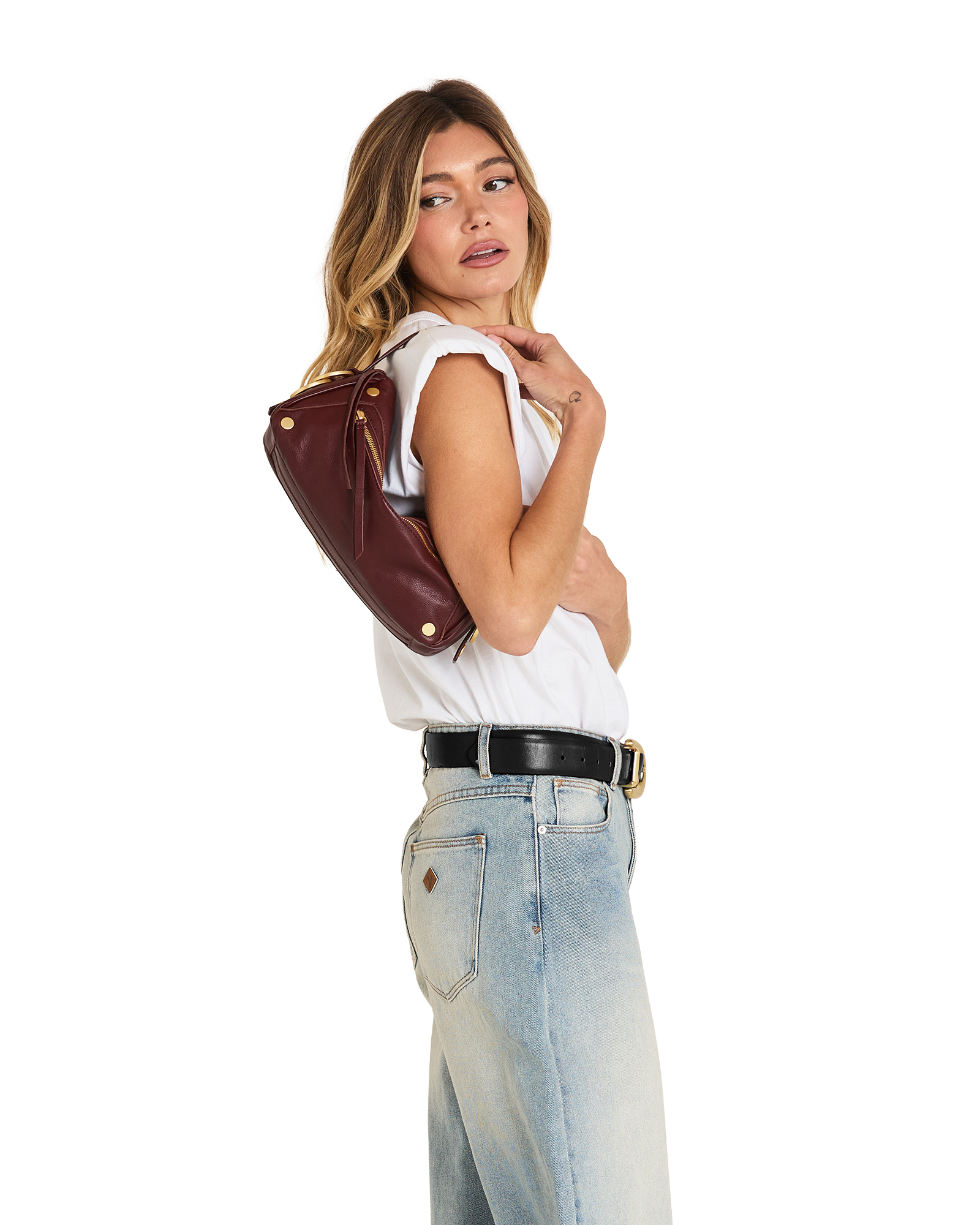 A woman posing in a white sleeveless top, light wash jeans, and a black belt, carrying a dark brown leather shoulder bag with gold-tone hardware against a plain white background.