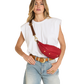 A woman modeling a red leather crossbody fanny pack with gold studs and a brown strap, styled with a white sleeveless top and light blue jeans against a white background.