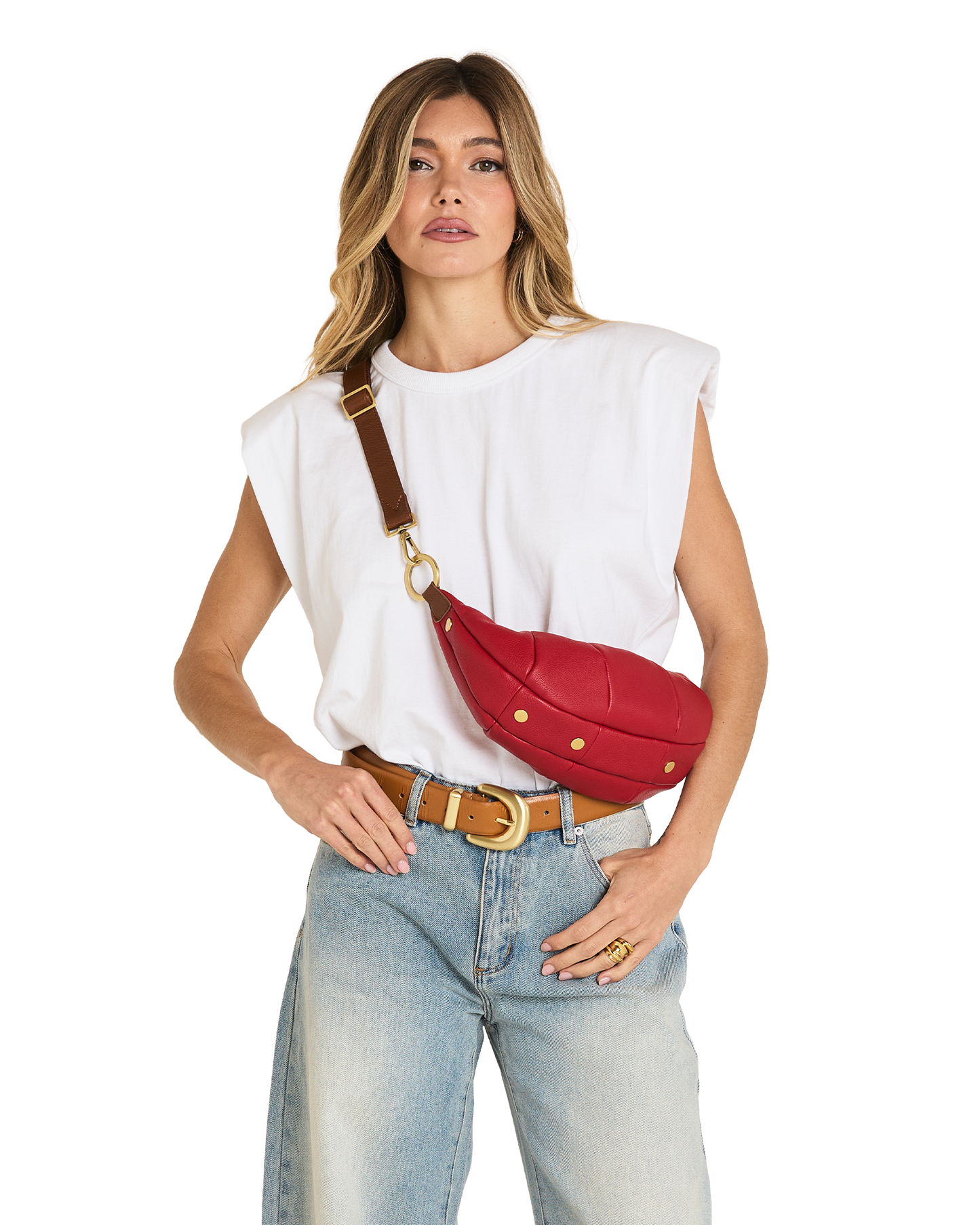 A woman modeling a red leather crossbody fanny pack with gold studs and a brown strap, styled with a white sleeveless top and light blue jeans against a white background.
