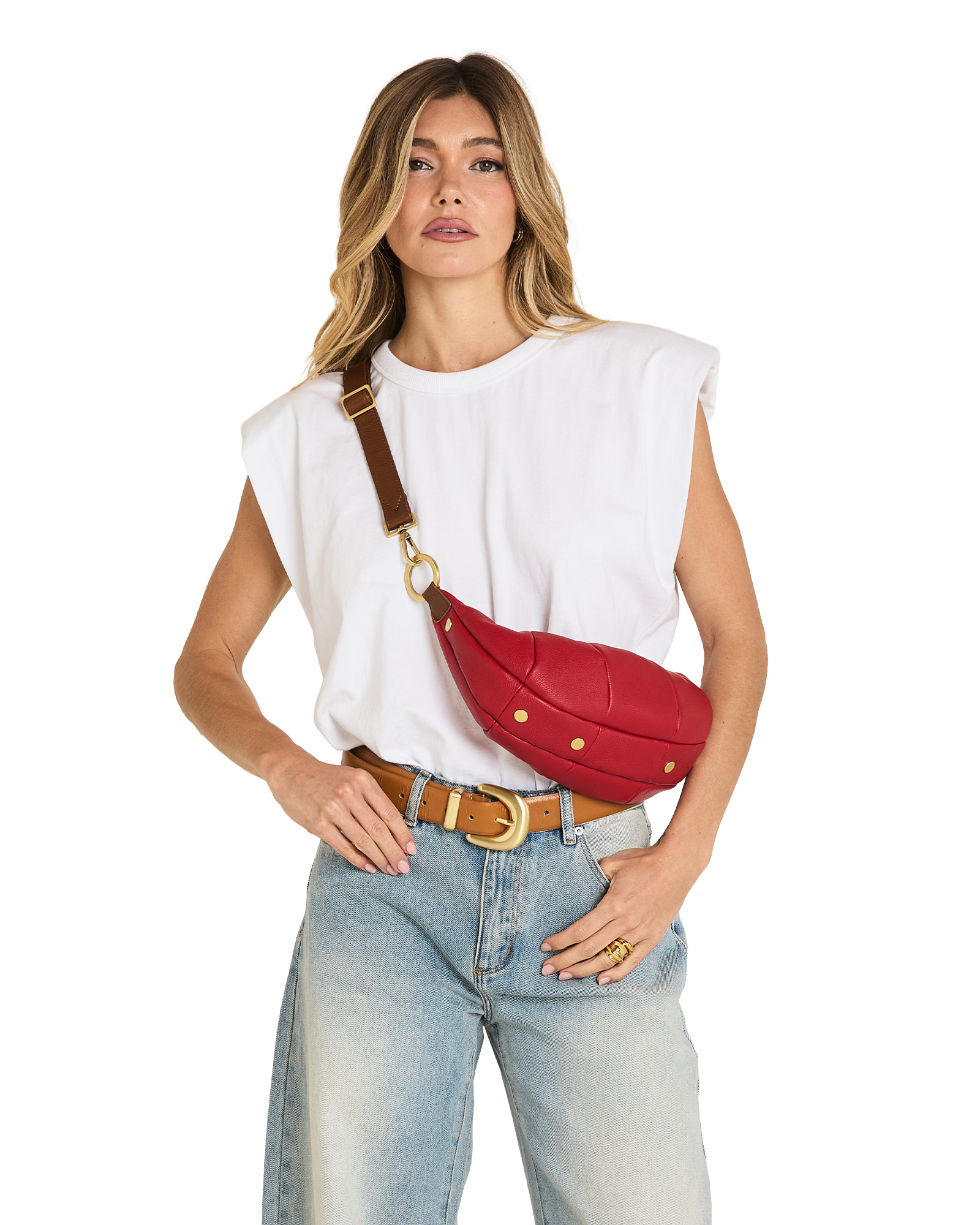 A woman modeling a red leather crossbody fanny pack with gold studs and a brown strap, styled with a white sleeveless top and light blue jeans against a white background.
