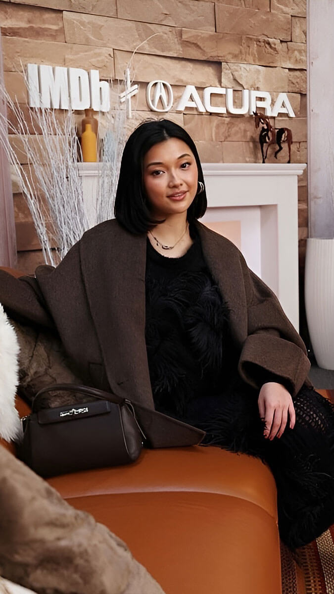 A woman sits on a brown leather sofa in a styled indoor setting, wearing a dark brown coat over a black outfit. A black handbag rests on the sofa beside her.